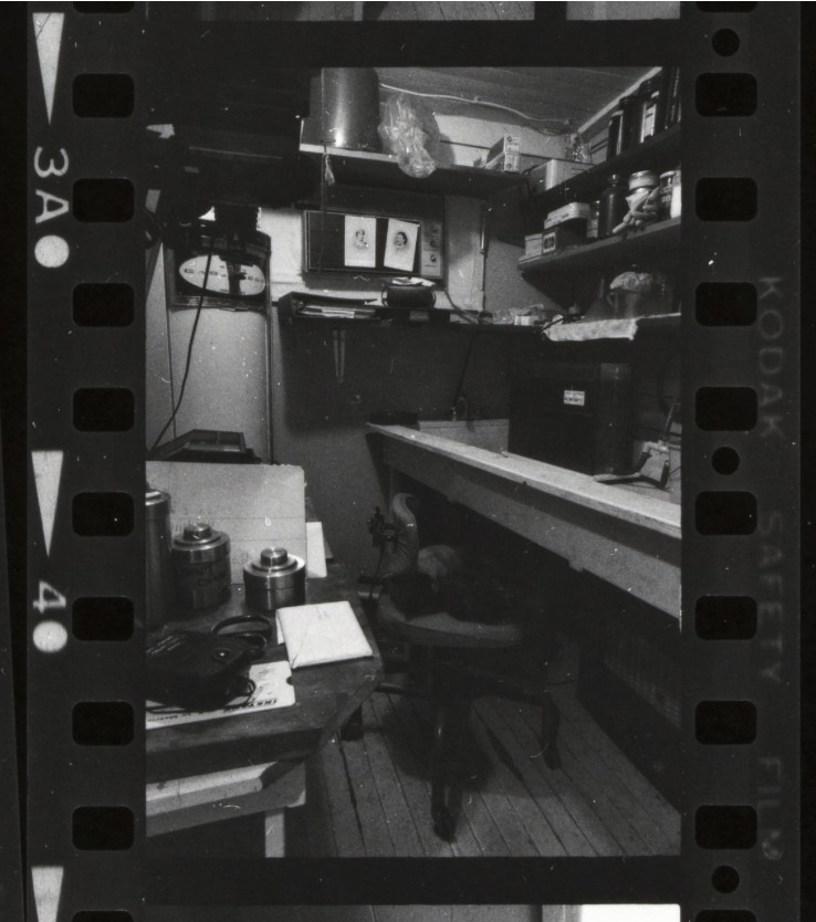 Black-and-white film frame of a small darkroom: a long wooden sink, an office chair, shelves of chemical bottles, a wall timer and film canisters on a side table.