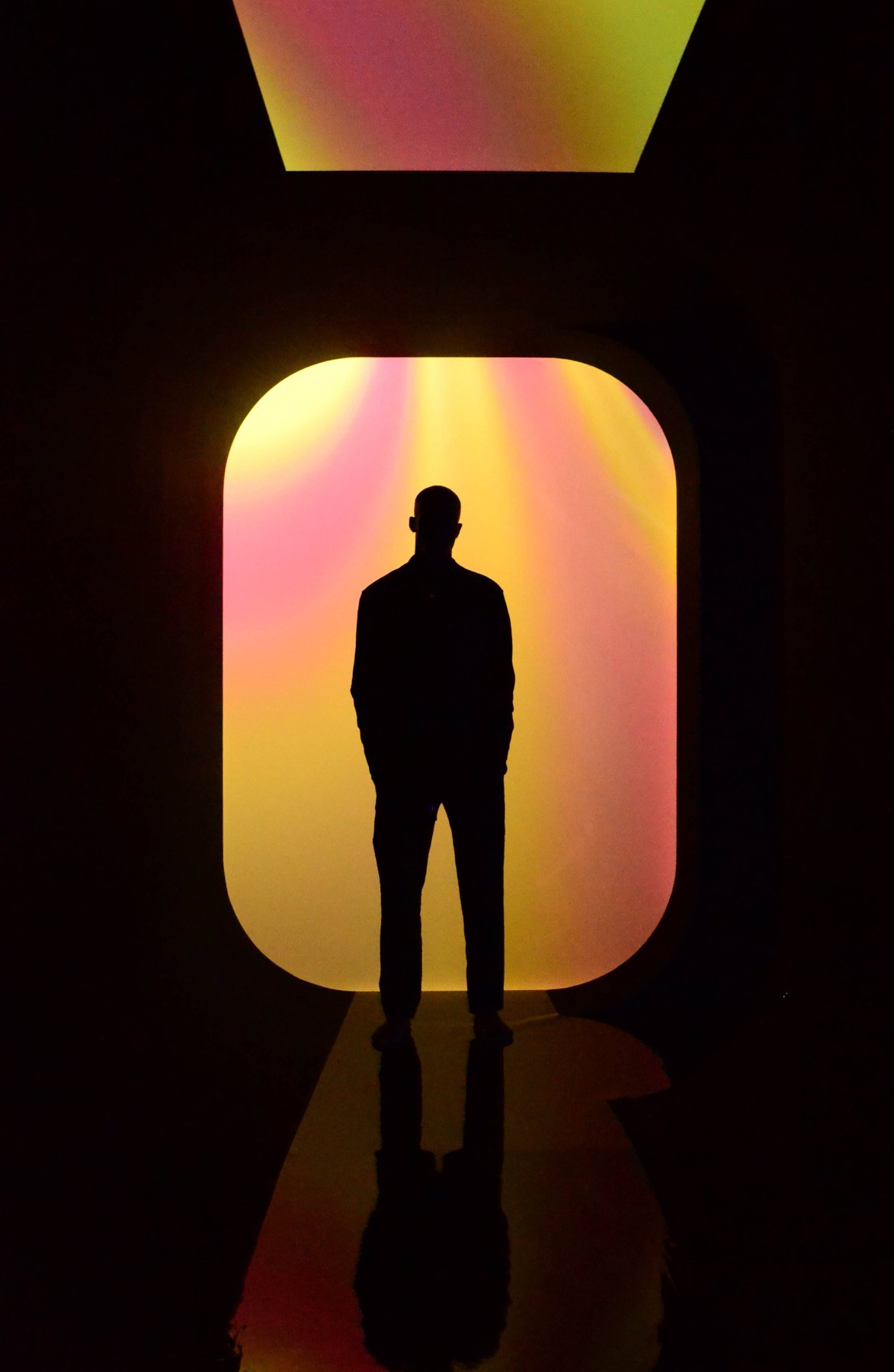 A silhouetted figure stands in a rounded doorway glowing yellow and pink, reflected on a glossy floor.