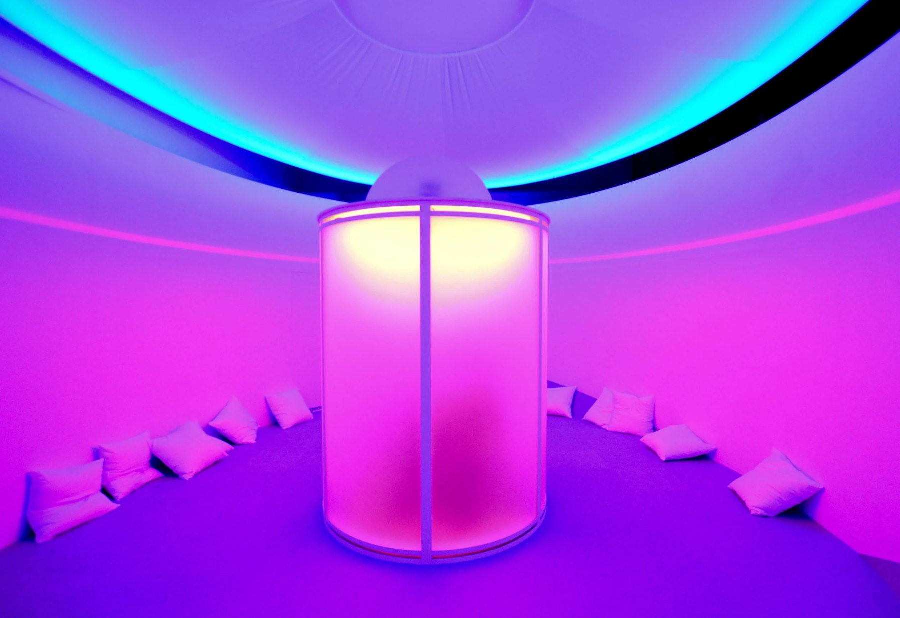 A round room lit violet and pink, with white cushions circling a glowing cylindrical chamber at the center under a ring of light.