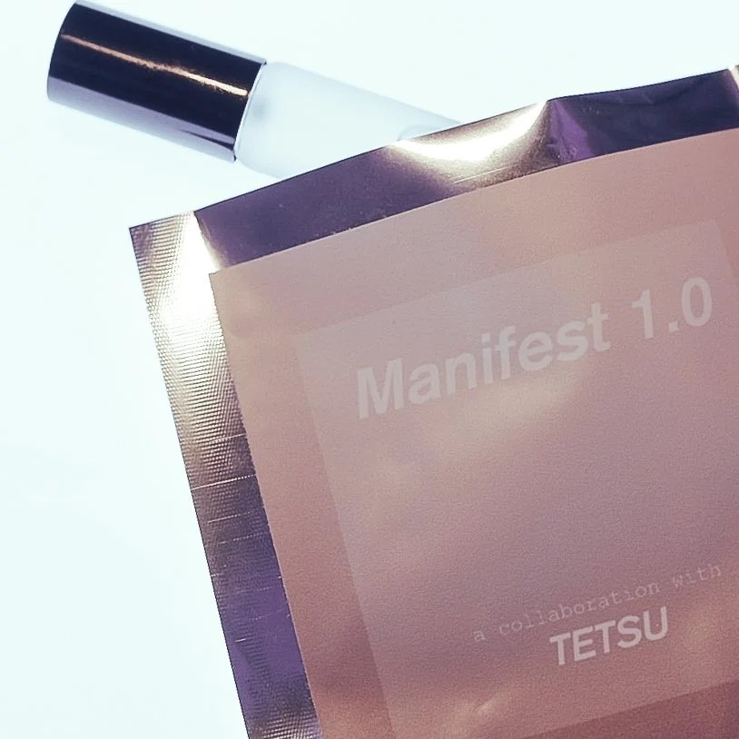 A frosted roller vial with a chrome cap beside a copper foil pouch printed Manifest 1.0, a collaboration with Tetsu.