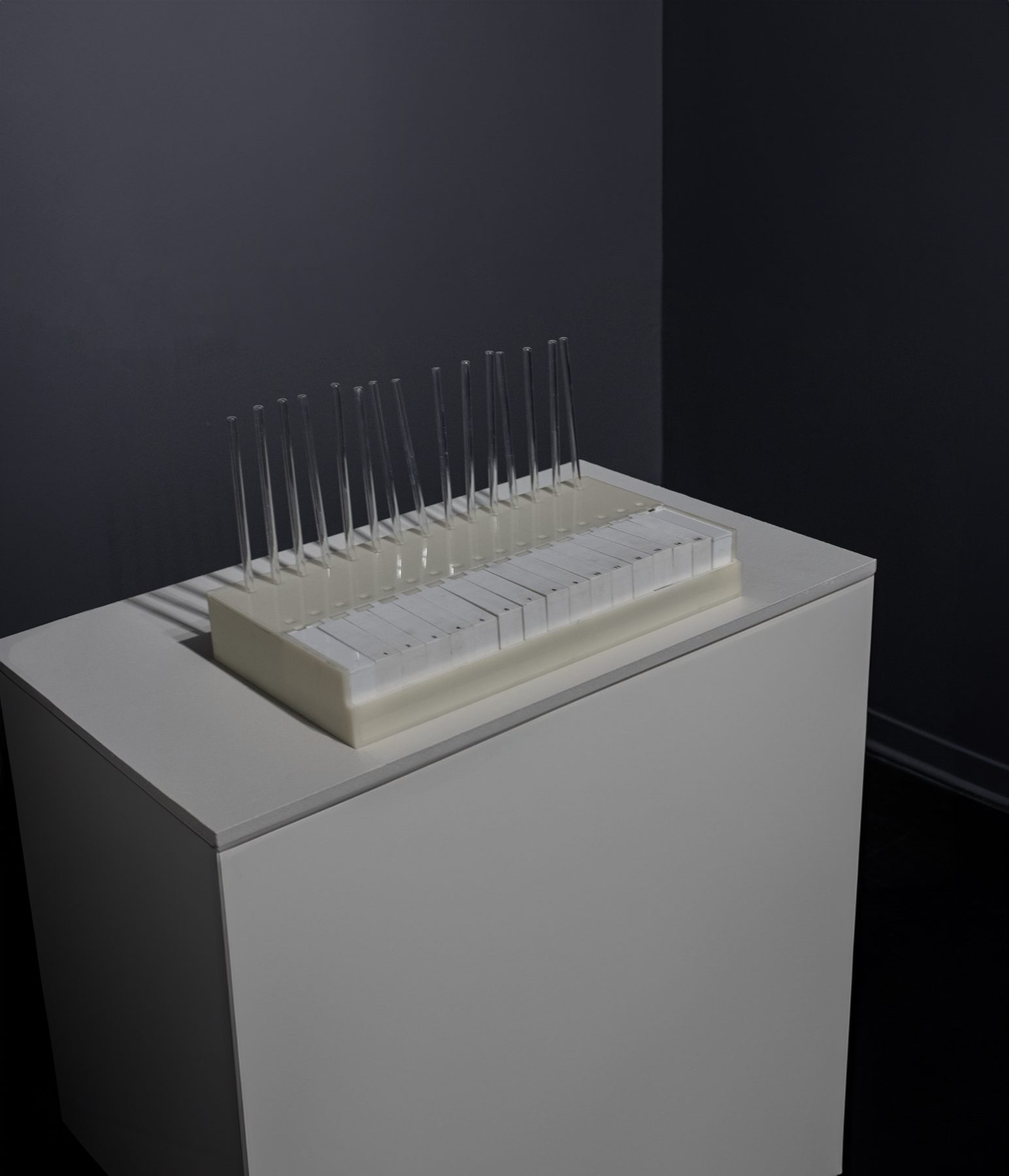 The Odor Organ on a white plinth in a dark gallery: a pale keyboard of fifteen keys with fifteen clear glass tubes rising behind it.