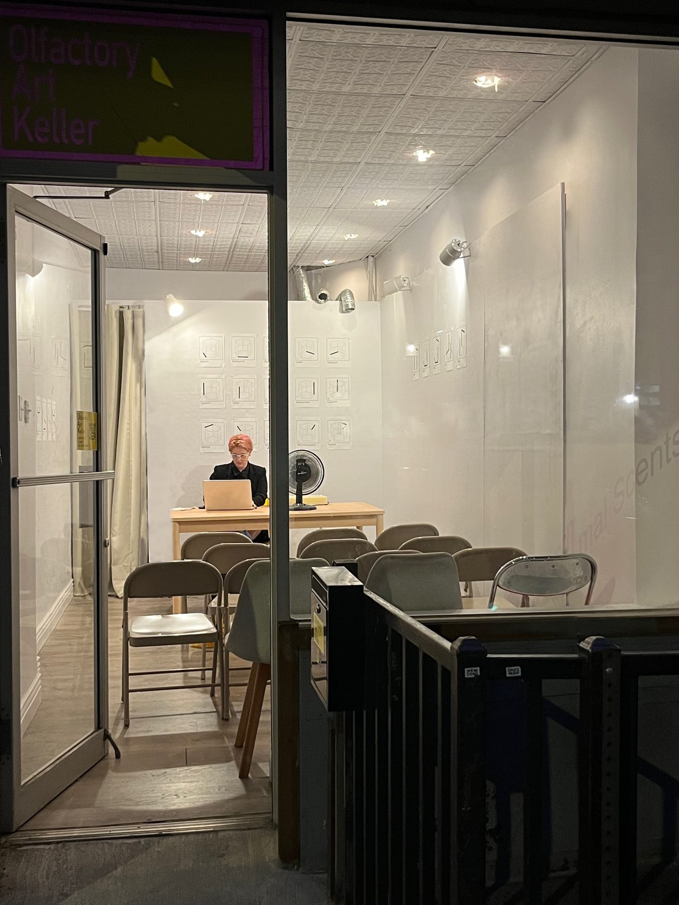 Night view through the window of Olfactory Art Keller before Symphonic Ephemera: M Dougherty at a table with the Odor Organ, notation prints on the walls, chairs set out for the audience.