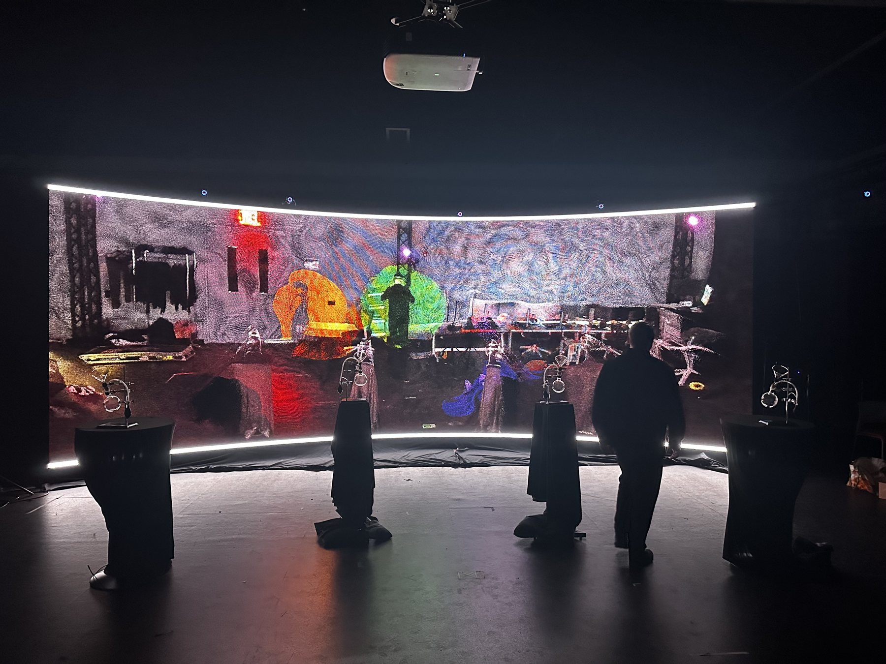 A curved LED wall shows a colorful mirrored scene of the room, with four pillars with diffusers stand in front of it in the dark.
