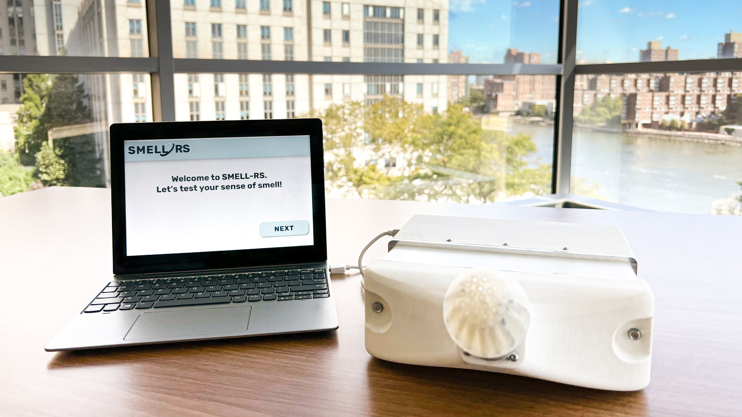A laptop showing the SMELL-RS welcome screen beside the white SMELL-RS device with its round sniffing port, on a table by a window overlooking a river.