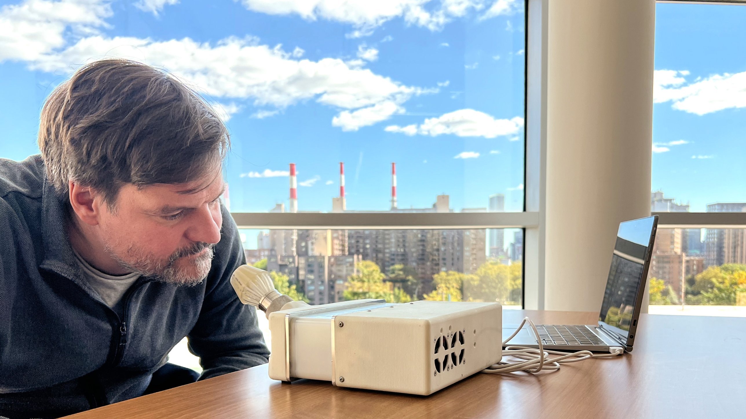Dr. Andreas Keller leans in to sniff from the white cone-shaped port on the side of the SMELL-RS device, a laptop beside it, at a window with a city skyline behind.